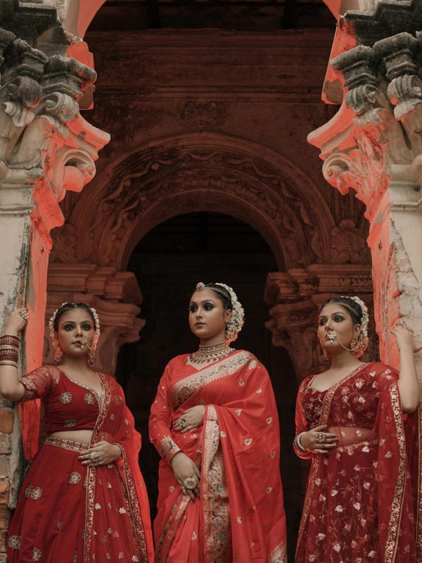 Three women in vibrant red sarees pose elegantly by ancient architecture.